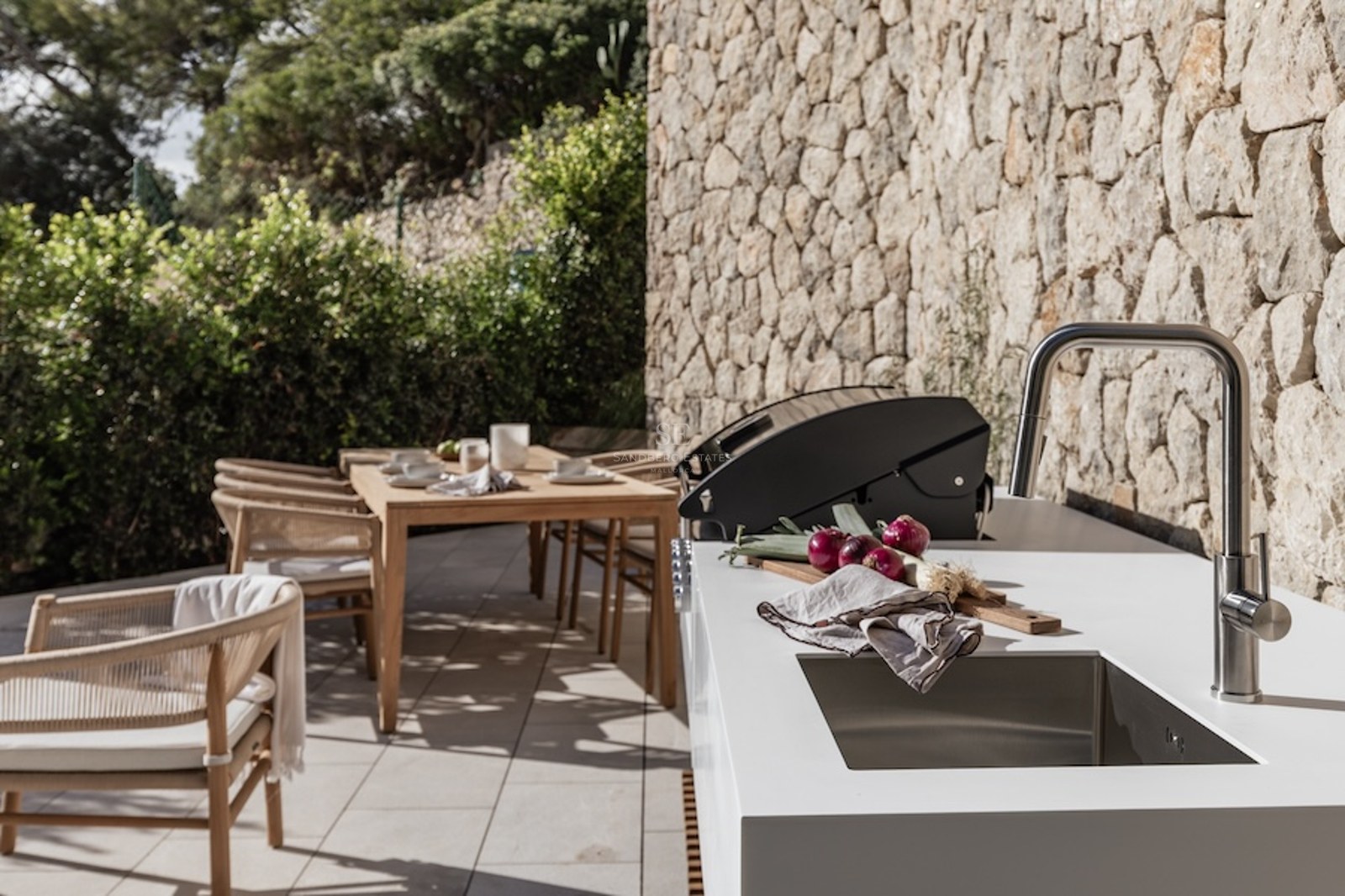 Modern outdoor kitchen with white countertop, sink, and grill next to a stone wall and wooden dining set.