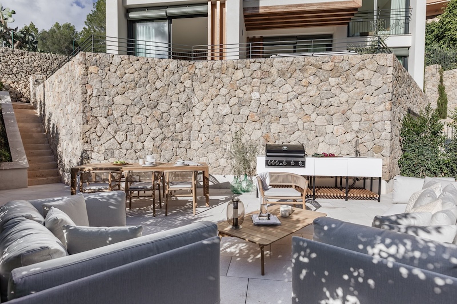 Modern terrace with a stone wall, outdoor kitchen, wooden dining table, and grey sofas under dappled natural light.