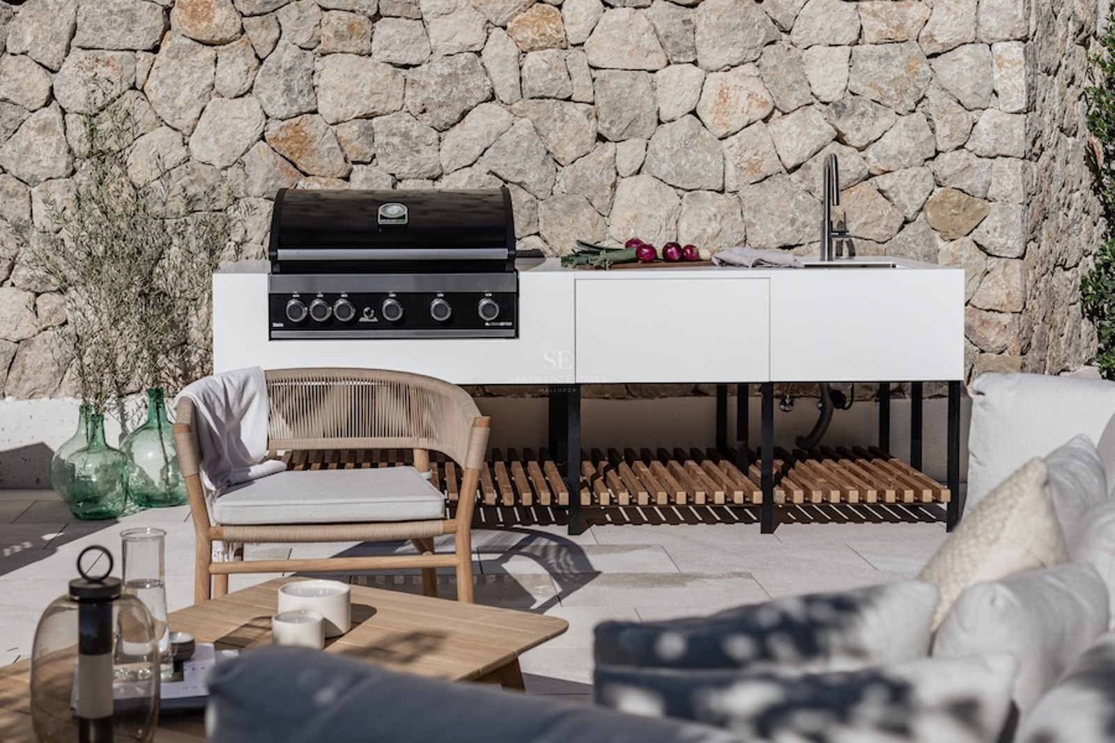Modern white outdoor kitchen with gas grill and sink against a stone wall with designer patio furniture.