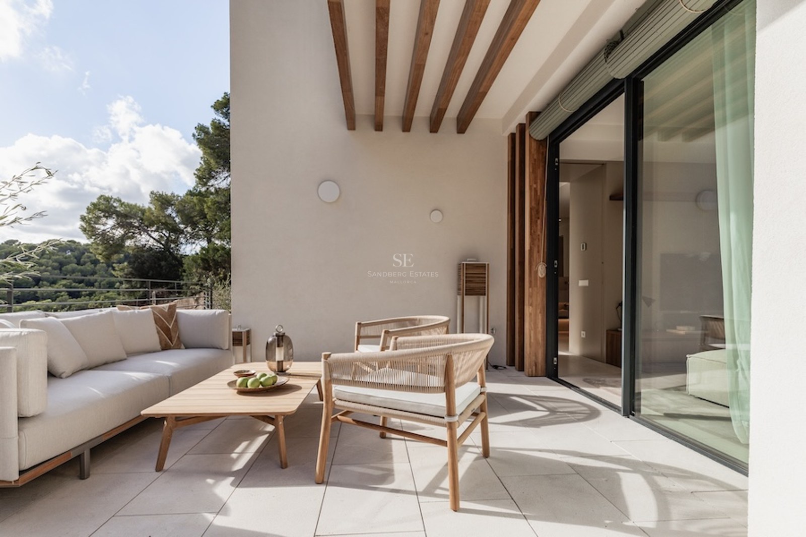 A sunlit modern terrace with a white sofa, wooden furniture, and views of lush green trees through glass doors.
