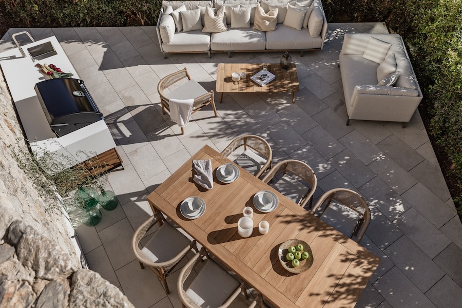 Overhead view of a modern stone patio featuring a teak dining set, white sofas, and a sleek outdoor kitchen with BBQ.