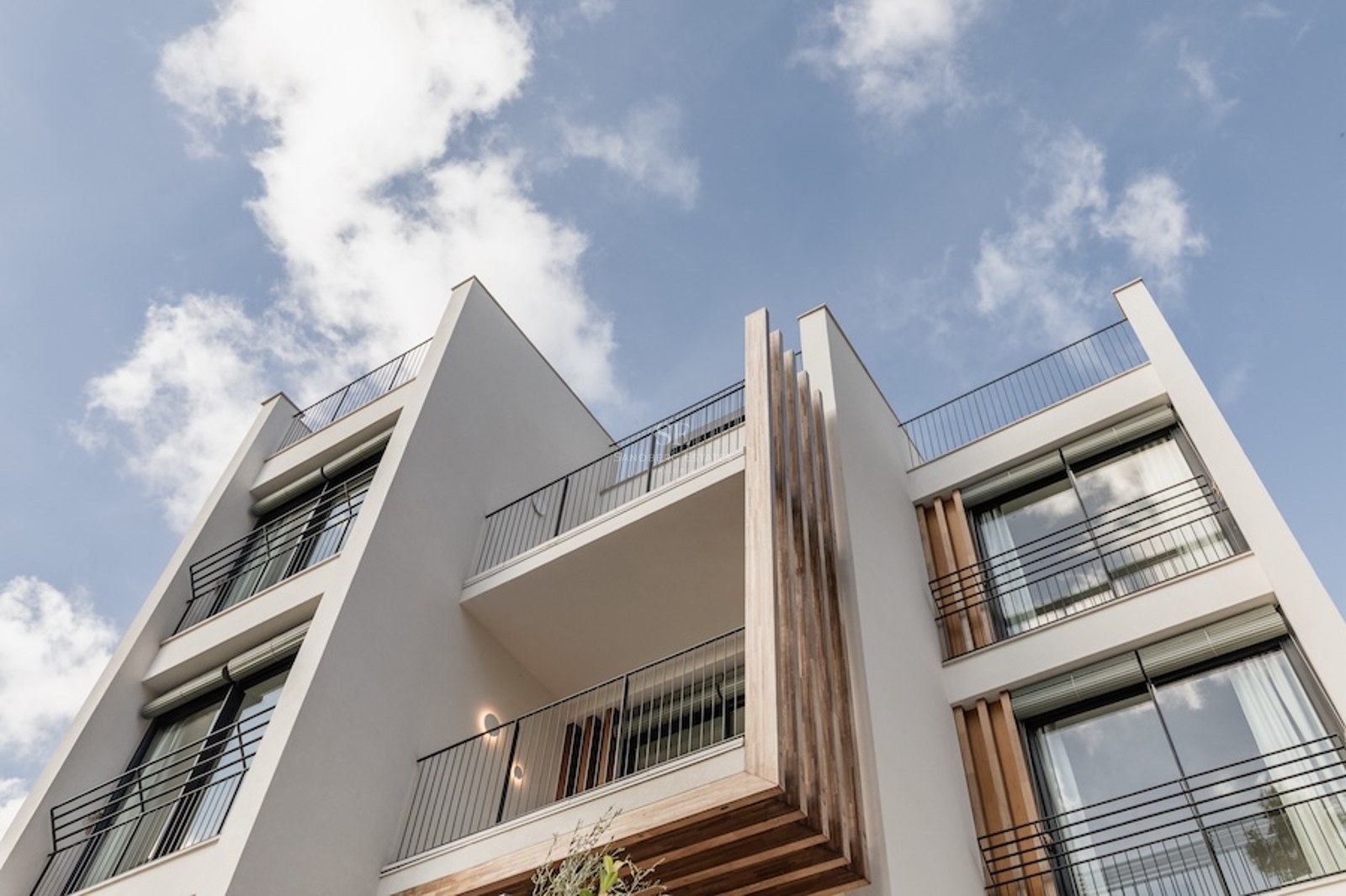Low-angle view of a modern white building facade with wooden accents and black balcony railings against a blue sky.