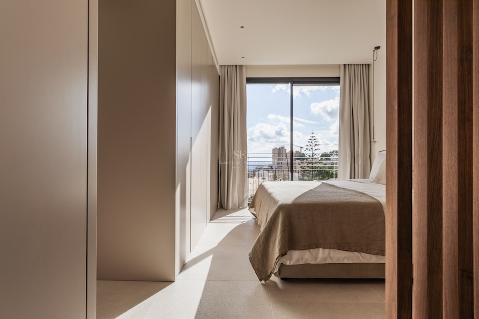 Modern bedroom with beige bedding, built-in wardrobes, and a large sliding glass door opening to a city balcony.