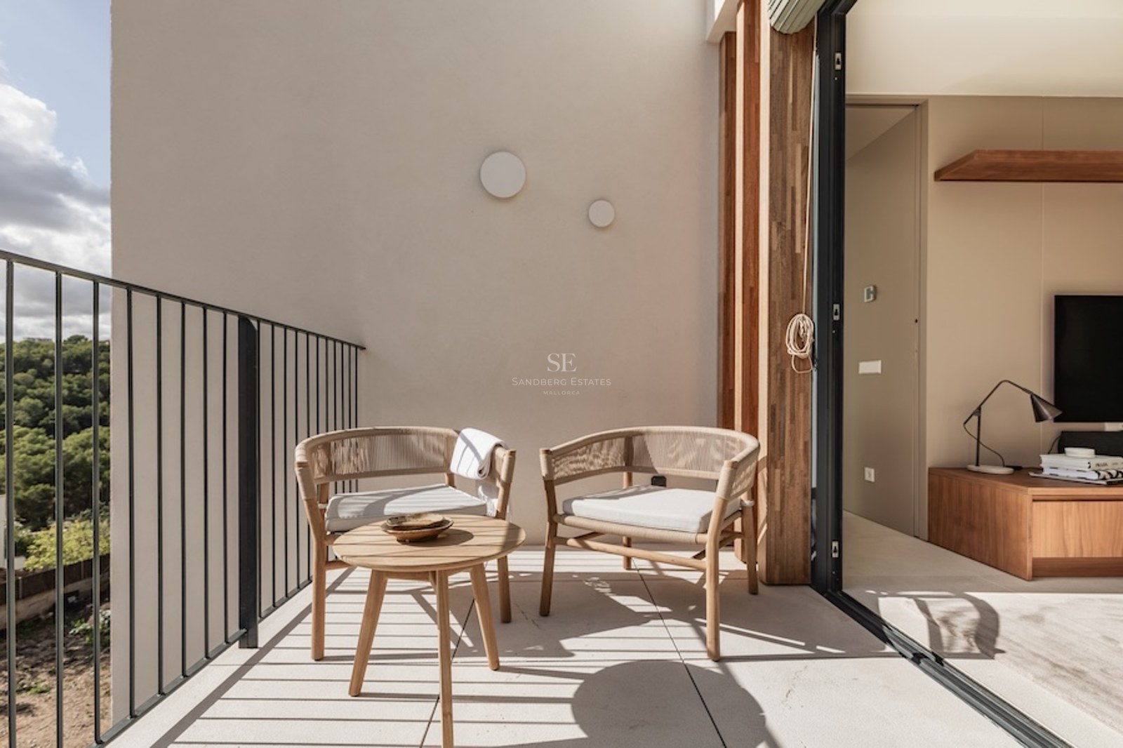 Sunlit balcony with designer woven chairs and a wooden coffee table overlooking a lush green forest.