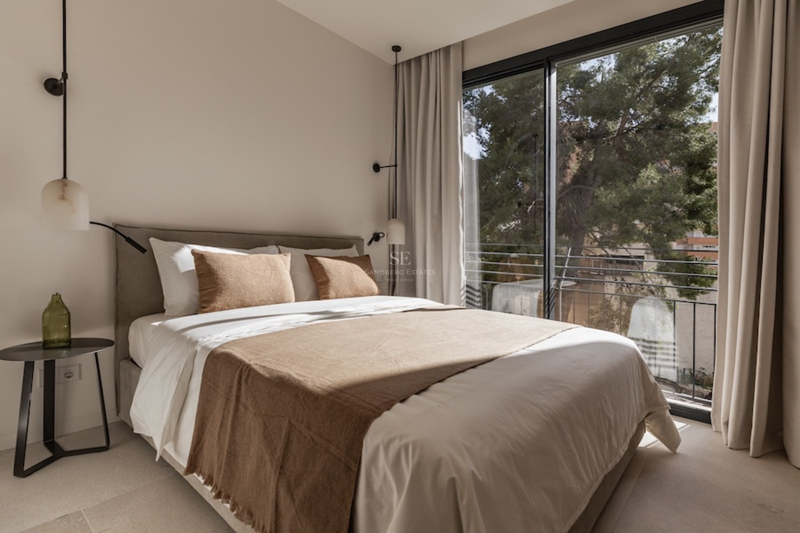 Bright bedroom with a large bed, neutral linens, designer pendant lights, and a floor-to-ceiling window viewing trees.