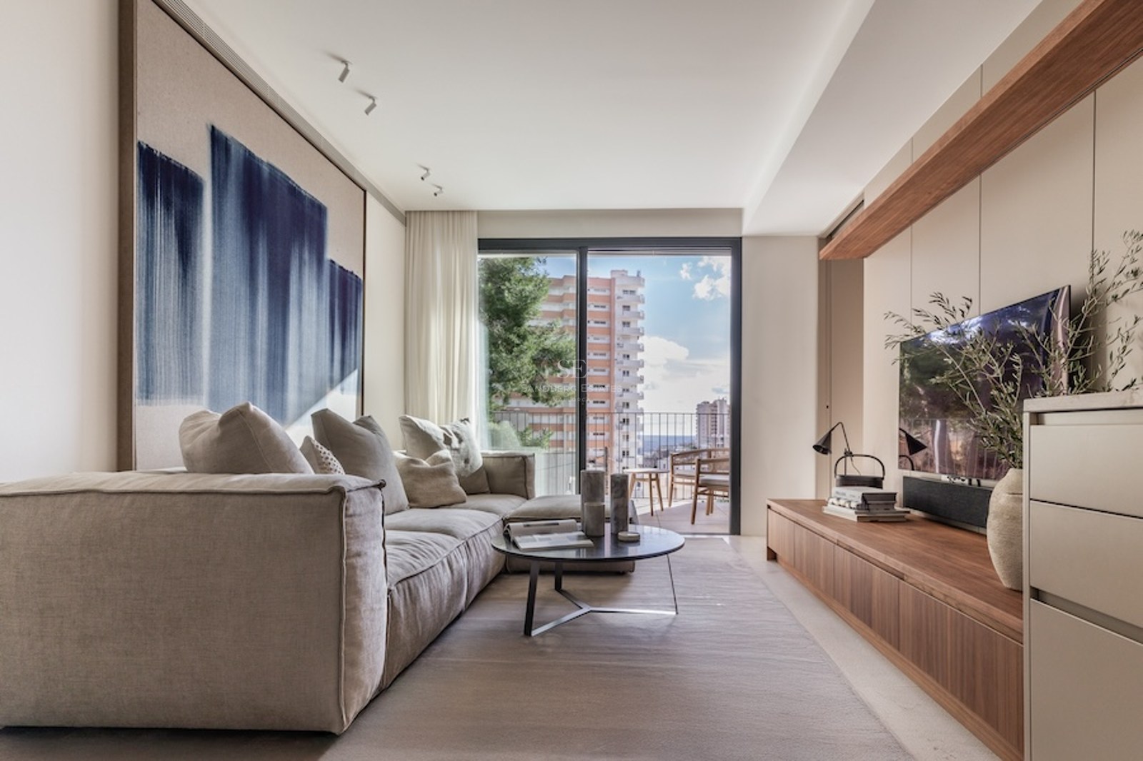 Bright modern living room featuring a plush beige sofa, abstract blue art, wooden TV console, and a balcony view.