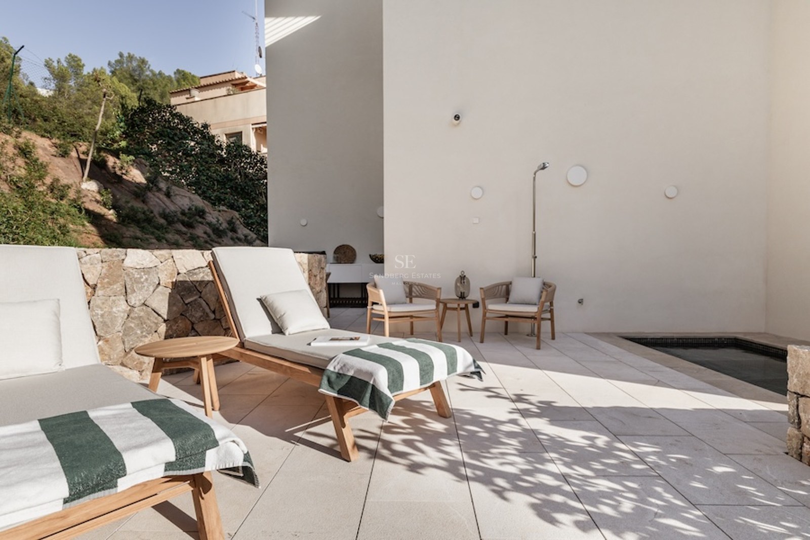 Modern outdoor terrace featuring wooden sunbeds, a small plunge pool, and a natural stone wall in bright sunlight.
