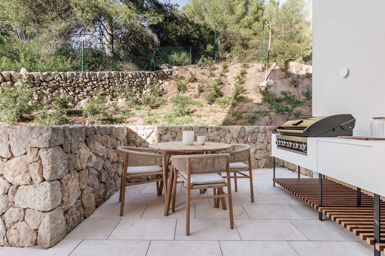 Outdoor terrace featuring a wooden dining set, stone walls, and a modern built-in grill area against a backdrop of trees.