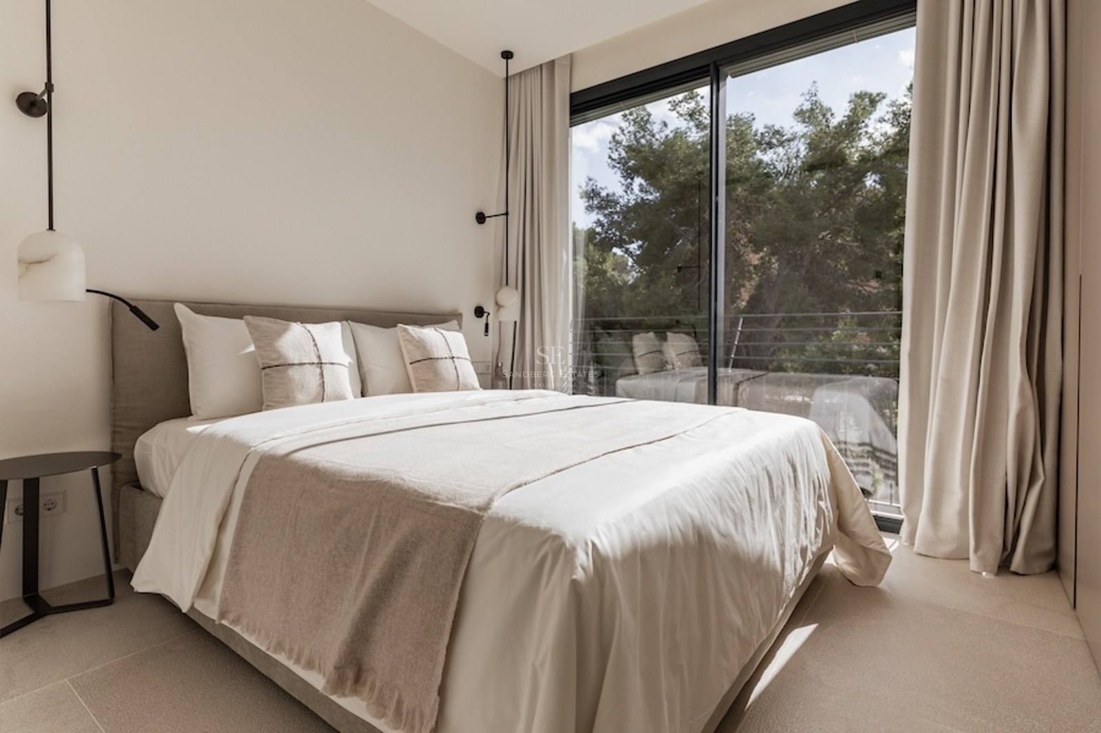 Helles Schlafzimmer mit Bett in neutralen Tönen vor einem großen Fenster mit Blick auf Bäume.