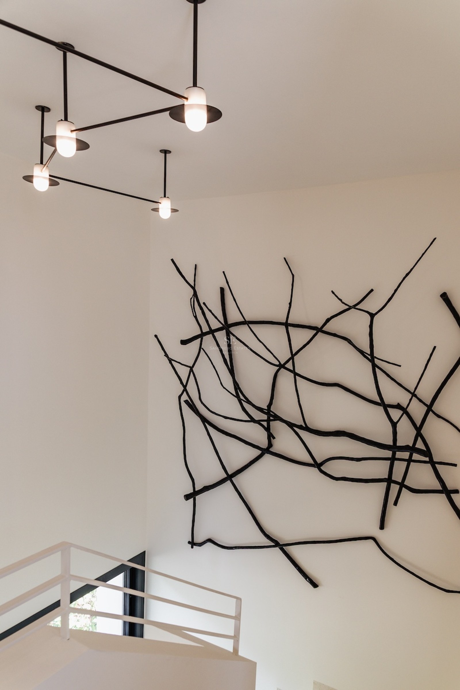 Schwarze, astähnliche Wandskulptur neben einem modernen Kronleuchter in einem weißen minimalistischen Raum mit Treppe.