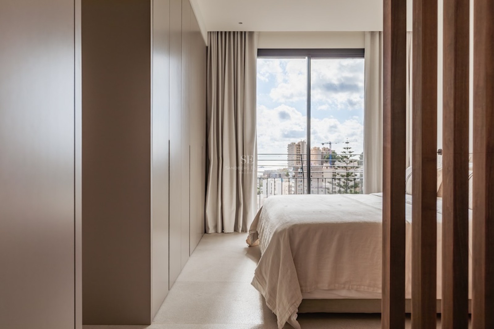 Helles Schlafzimmer in neutralen Tönen mit großem Einbauschrank und bodentiefem Fenster mit Blick auf die Stadt.