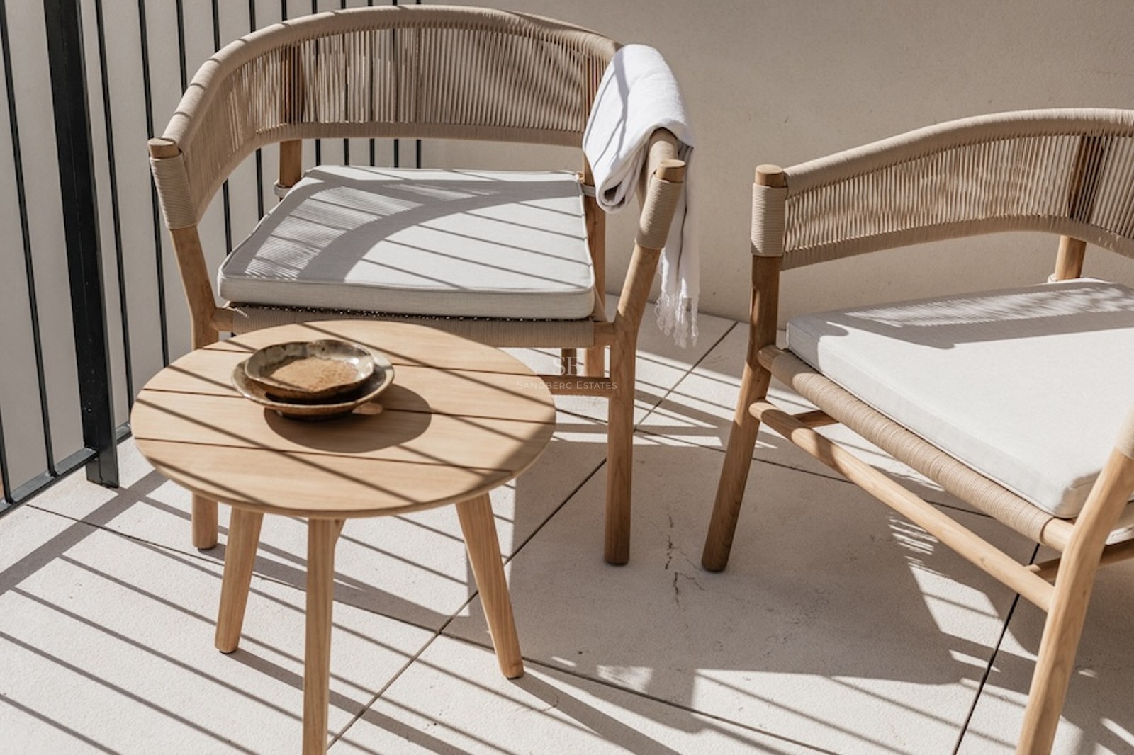 Zwei elegante geflochtene Stühle und ein runder Holzbeistelltisch auf einem sonnigen Balkon mit Schatten.