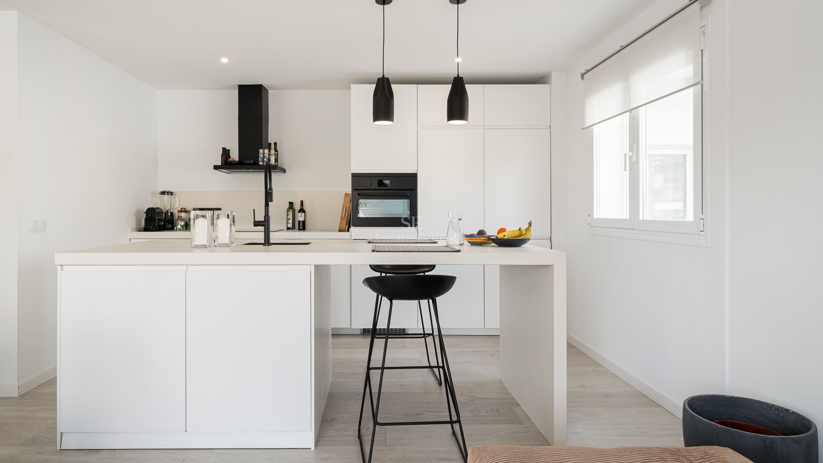 Bright white kitchen featuring a large central island with black bar stools and black hanging pendant lights.