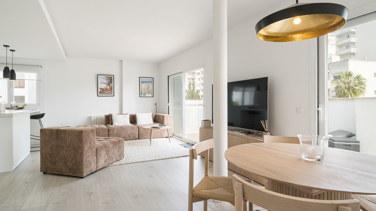 A sunlit living room featuring a brown modular sofa, wooden dining table, and large glass sliding doors leading to a balcony.