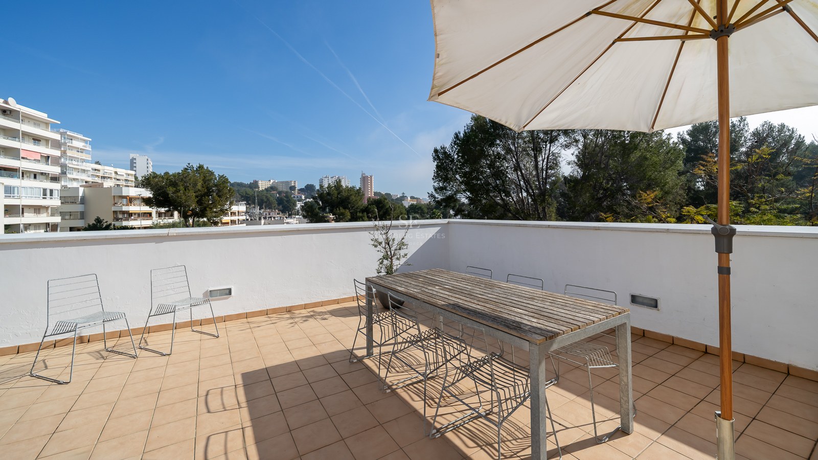 Spacious rooftop terrace with a large wooden table, wire chairs, and a white sun umbrella overlooking a city.