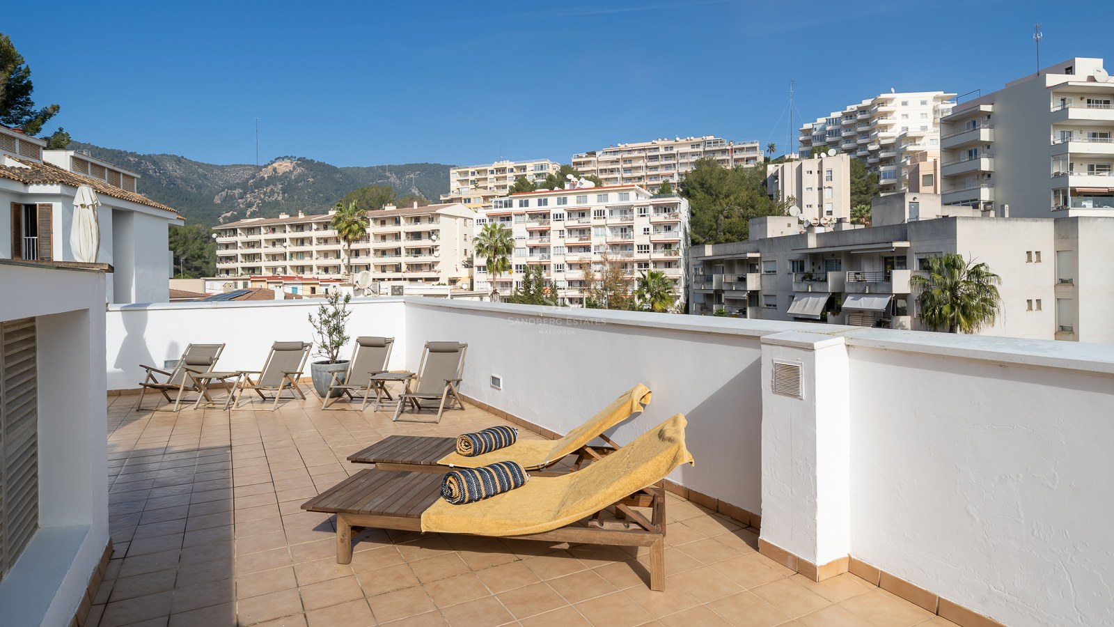 Spacious rooftop terrace featuring wooden sun loungers, tiled flooring, and views of urban buildings and mountains.