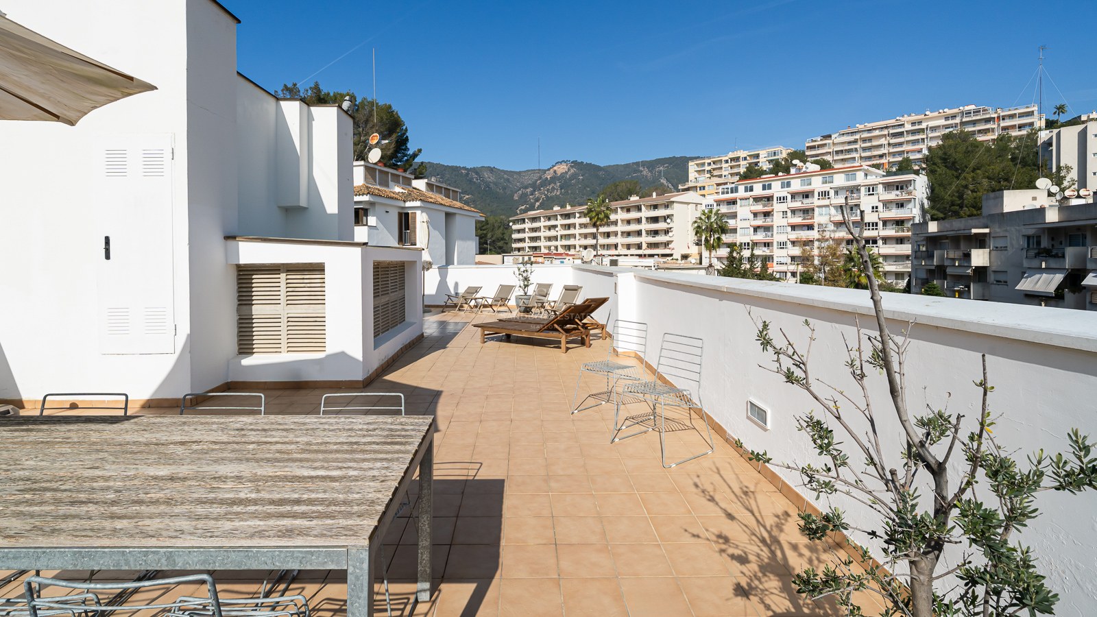 Large terrace with terracotta tiles, wooden sun loungers, and dining table under a clear blue sky.