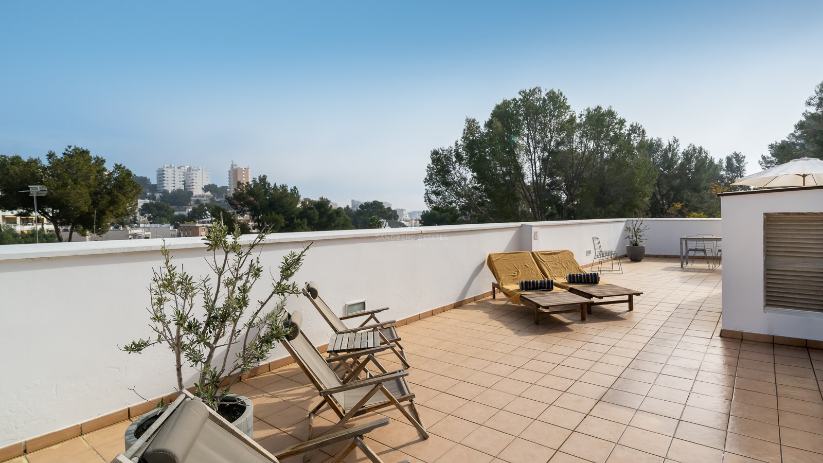 Large tiled terrace with wooden lounge chairs, potted plants, and a view of trees and city buildings.