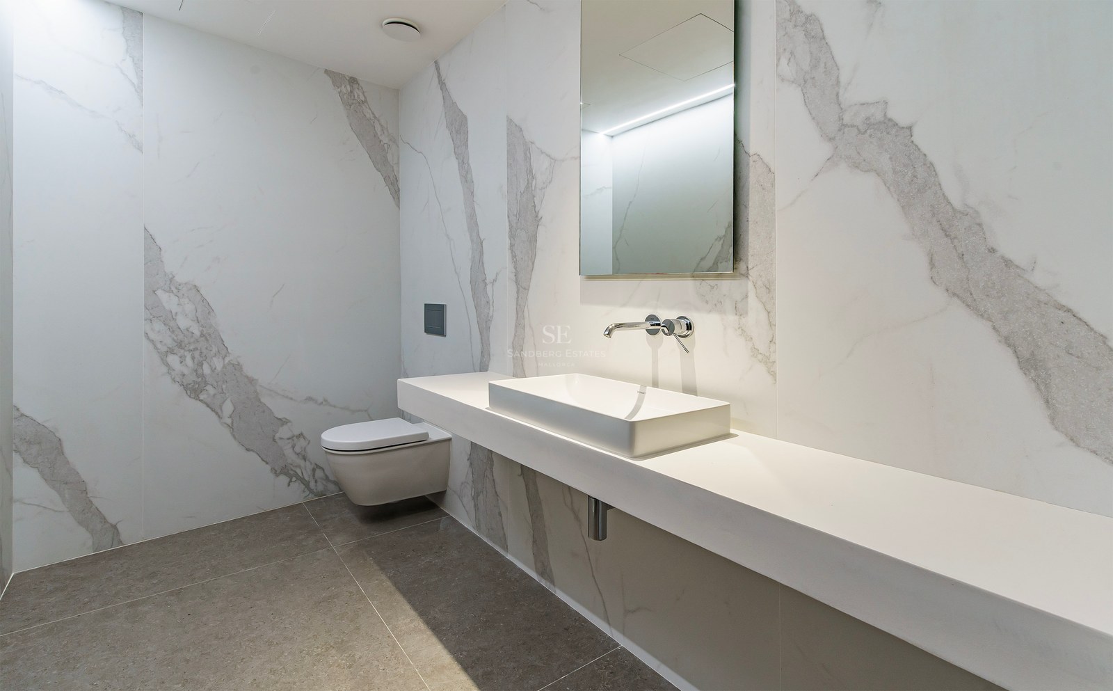 Baño moderno con revestimiento de mármol blanco, lavabo rectangular suspendido y grifería cromada empotrada.