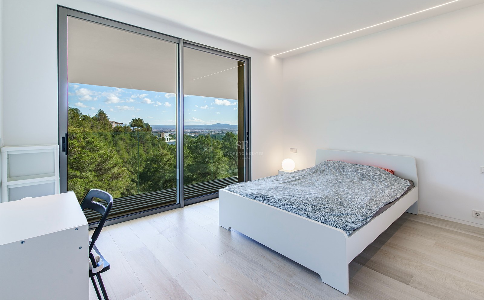 Dormitorio luminoso con cama blanca, suelo de madera clara y grandes ventanales con vistas a un bosque y montañas.
