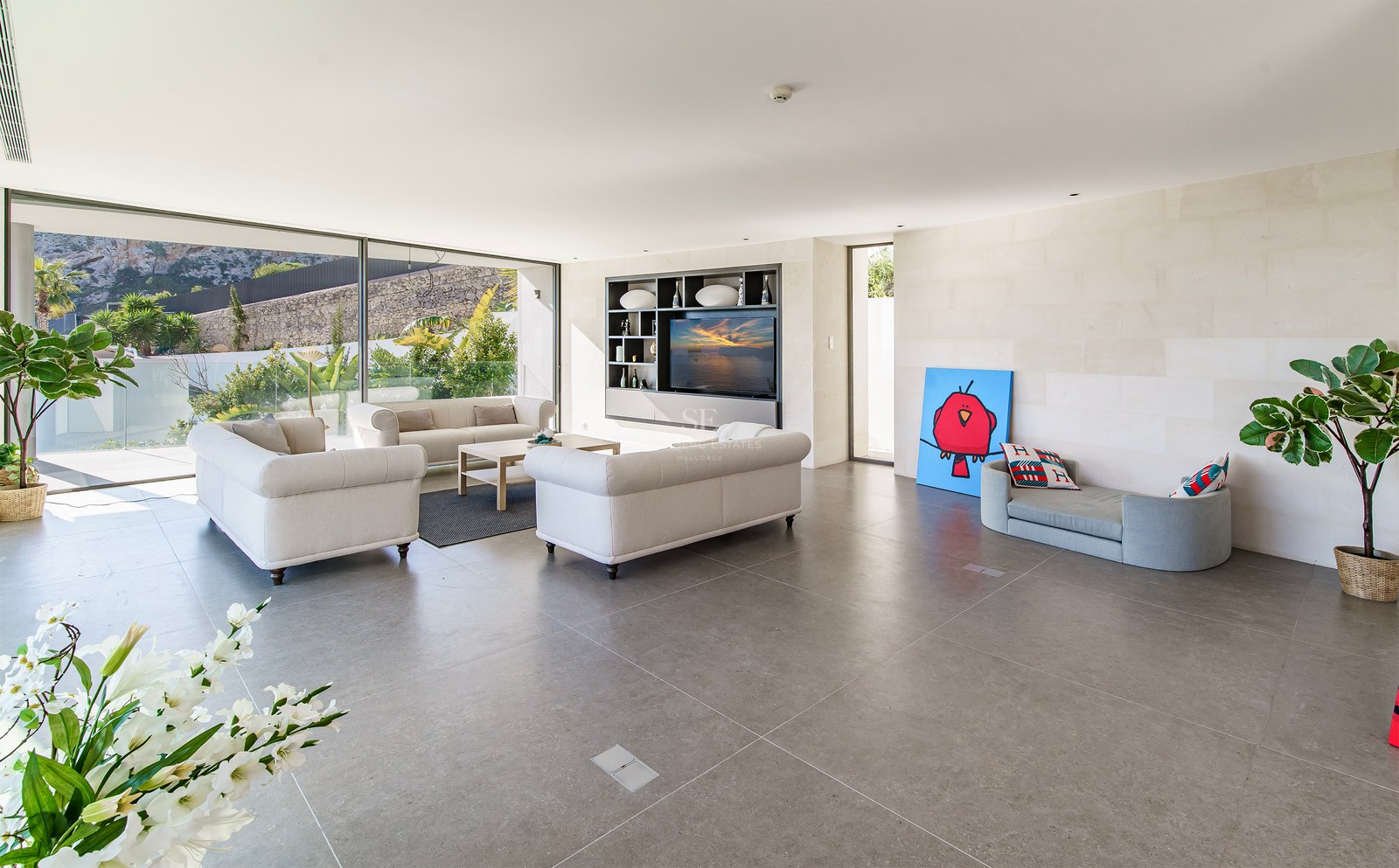 Salón moderno y luminoso con sofás beige, suelo de piedra gris y grandes ventanales hacia el jardín.