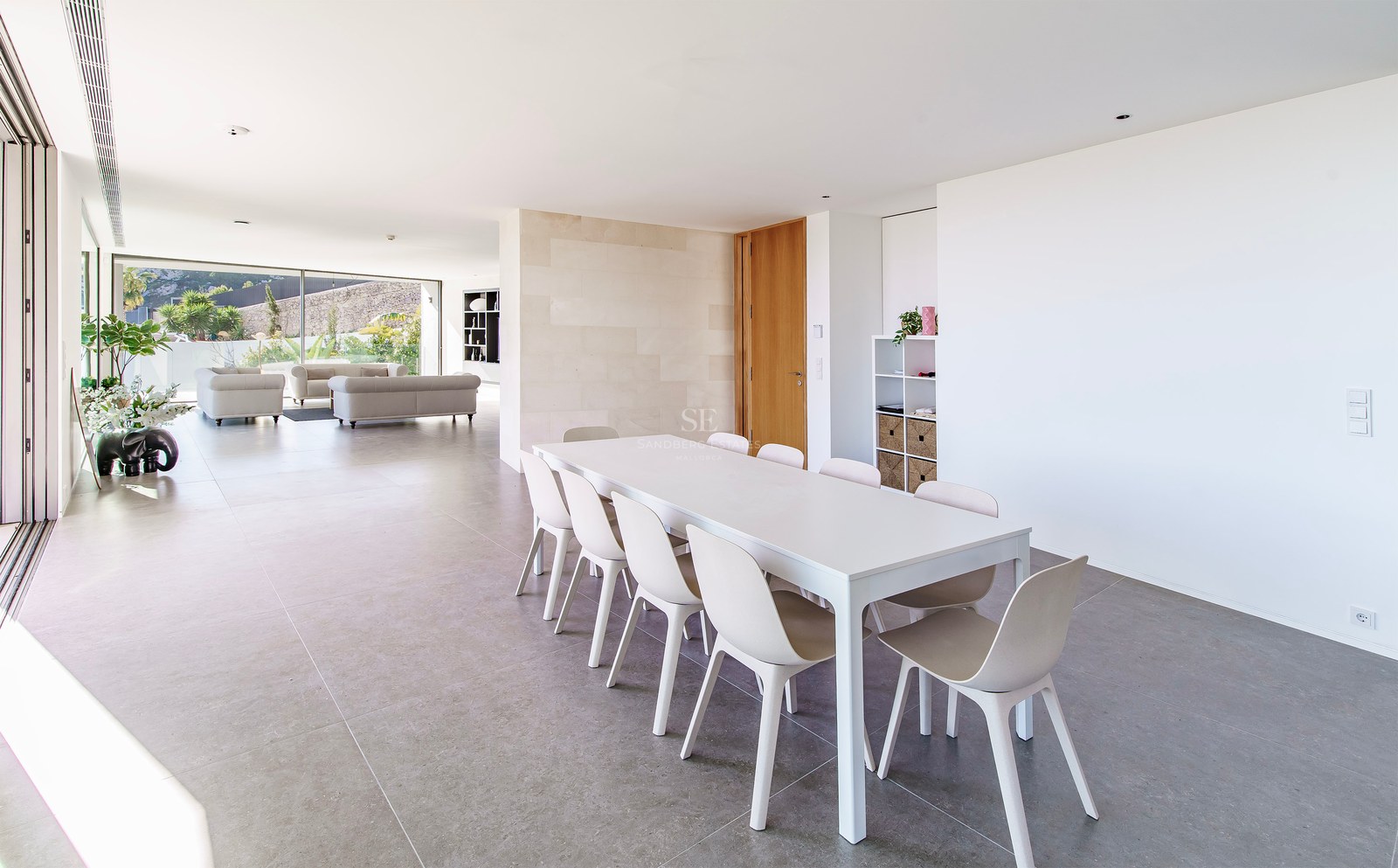 Comedor minimalista luminoso con mesa y sillas blancas, que da a una amplia sala de estar con ventanales.