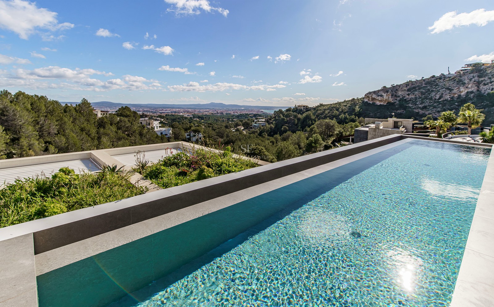 Piscina infinita con agua cristalina con vistas a un valle verde y una ciudad lejana bajo un cielo despejado.