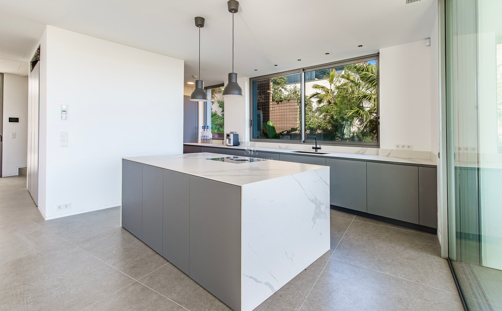 Cocina moderna con una gran isla de mármol blanco, gabinetes grises y ventana con vista a plantas tropicales.