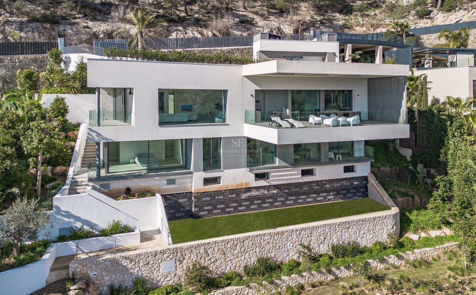 Villa moderna blanca de varias plantas con grandes ventanales y terrazas construida en una ladera rocosa.