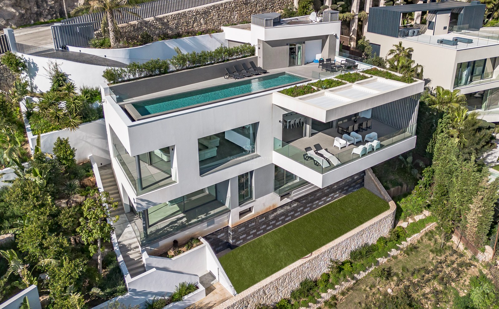Vista aérea de una villa blanca de tres niveles con piscina infinita en la azotea y terrazas de cristal.