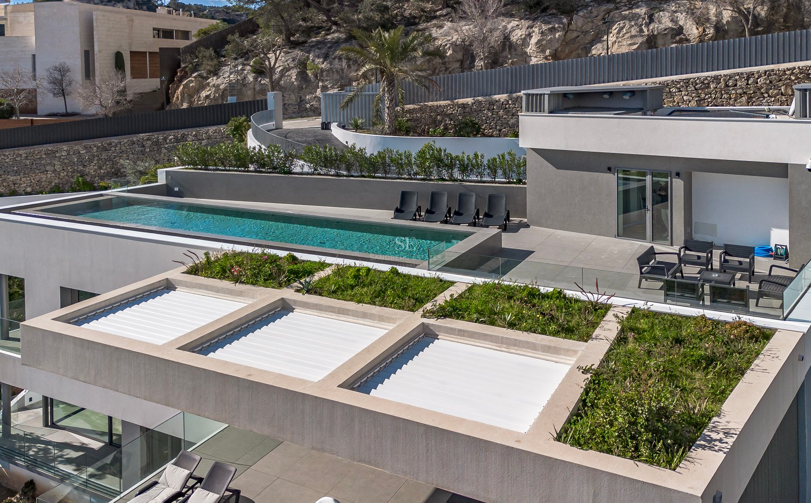 Vista aérea de una villa moderna gris con piscina infinita, tumbonas y vegetación en la azotea frente a una colina.