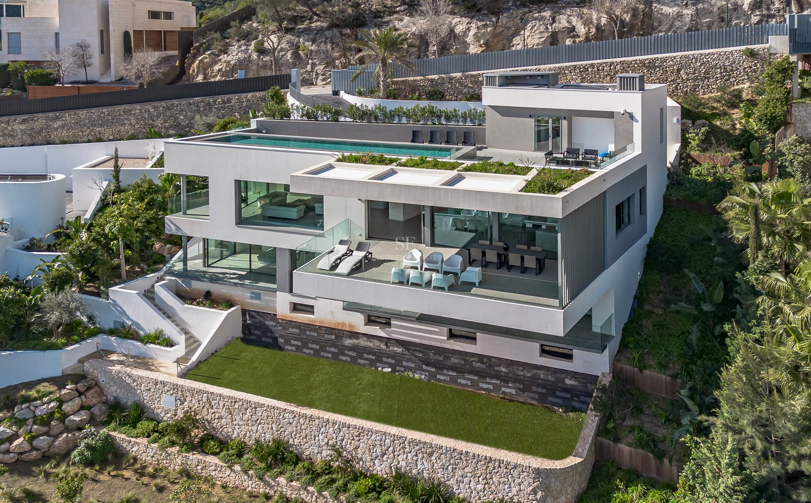 Vista aérea de una villa moderna blanca de varias plantas con piscina infinita en la azotea y terrazas de cristal.