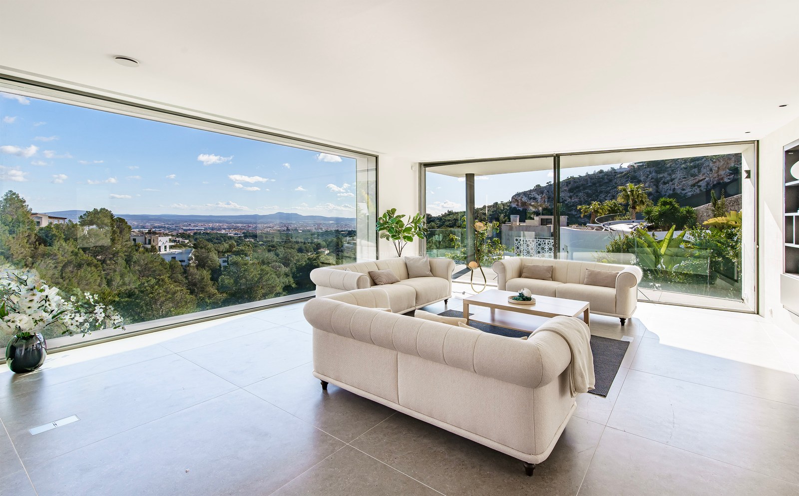 Salón moderno con tres sofás beige y grandes ventanales que muestran vistas a la ciudad y las montañas.
