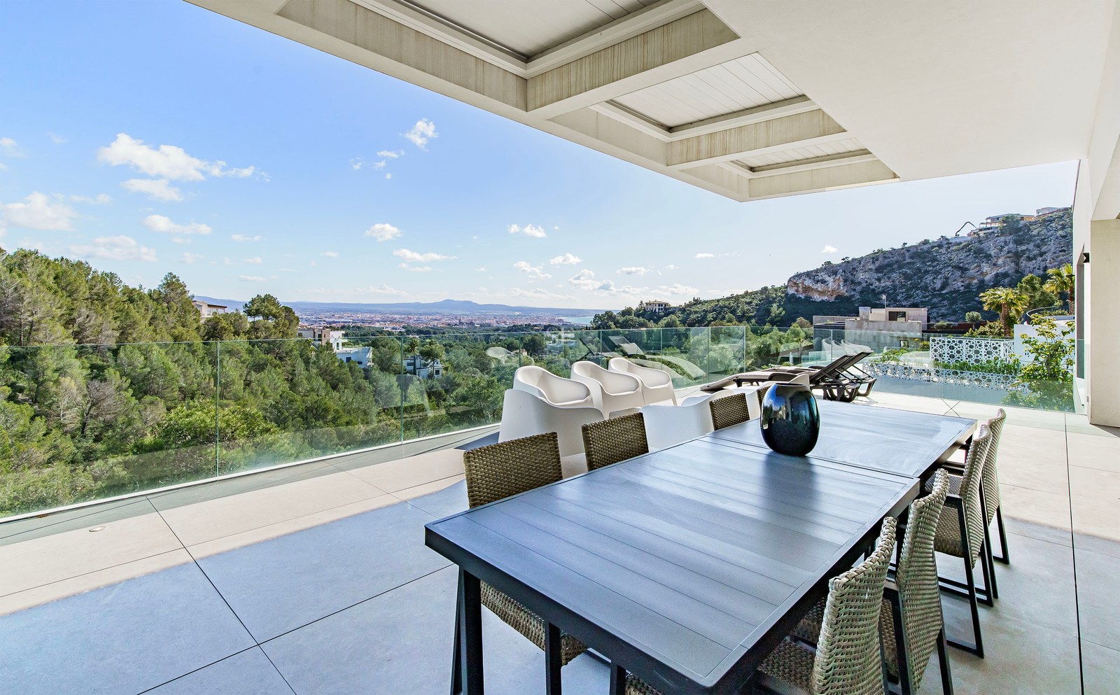 Terraza moderna con mesa de comedor y barandillas de cristal con vistas a un bosque y la ciudad lejana.