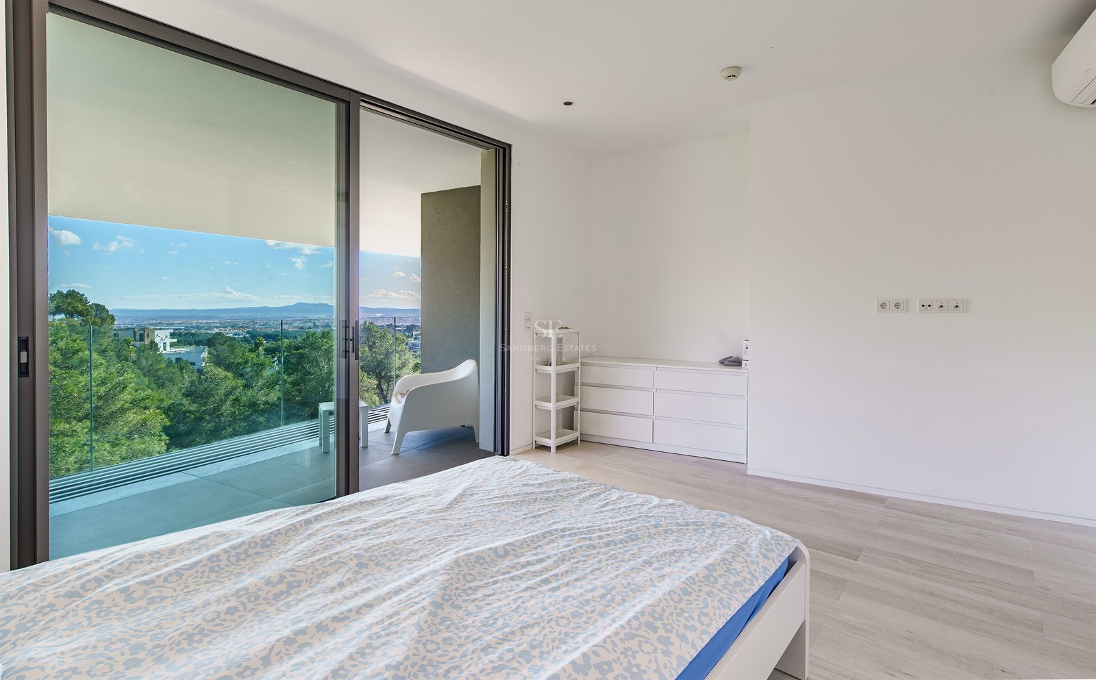 Dormitorio blanco minimalista con grandes puertas de cristal a una terraza con vistas al bosque.