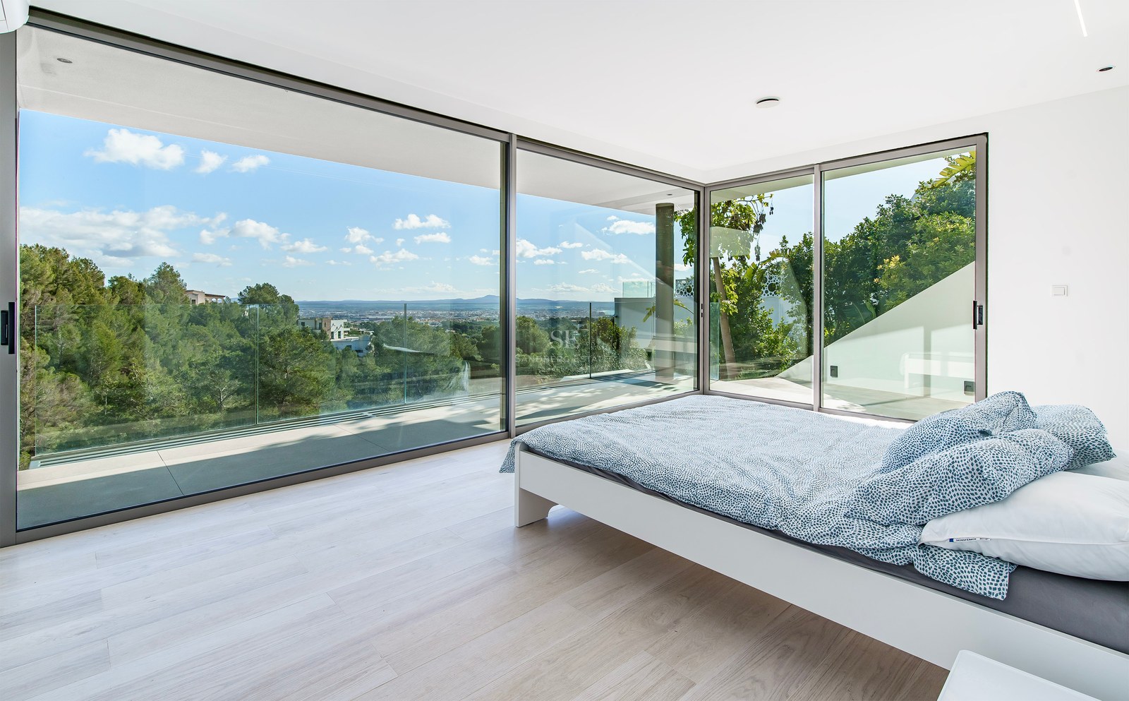 Dormitorio moderno con cama blanca y grandes ventanales con vistas a un bosque verde y la ciudad al fondo.