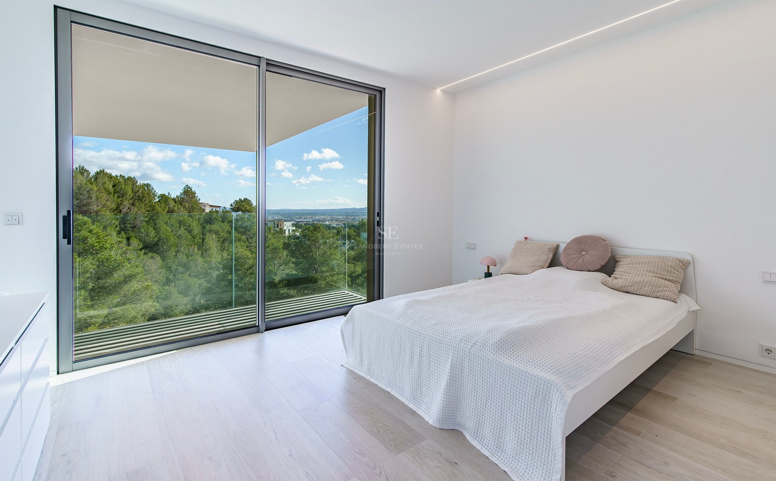 Dormitorio minimalista luminoso con grandes puertas correderas de cristal que dan a un balcón con vistas a colinas verdes.