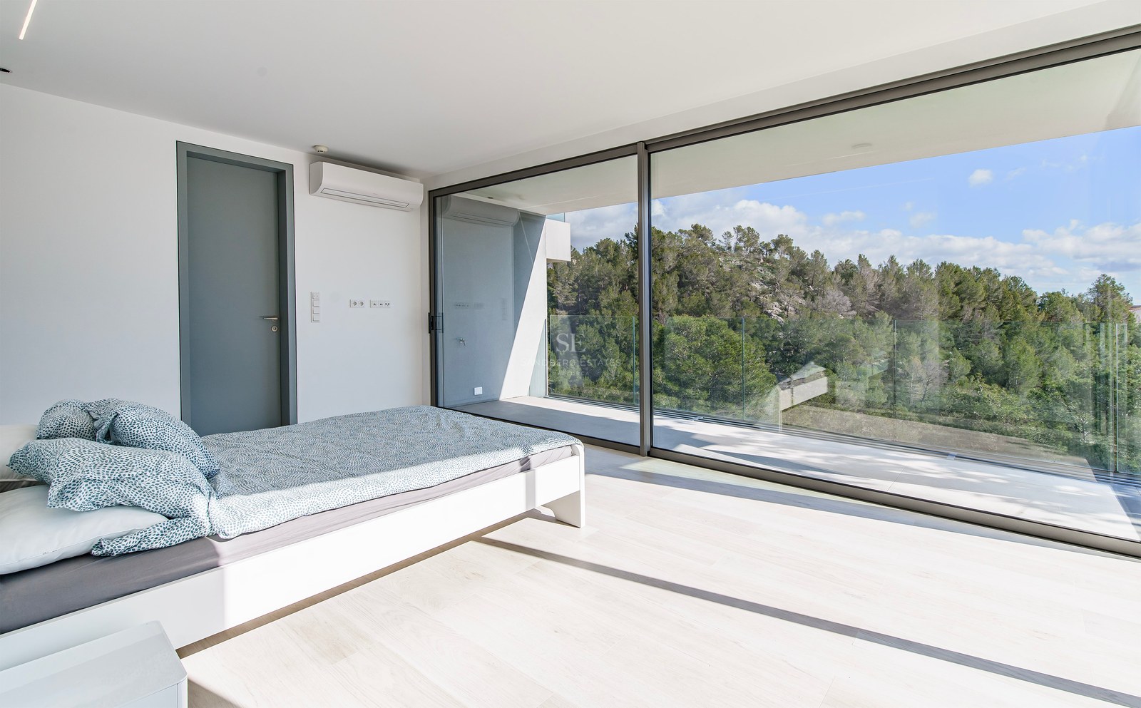 Dormitorio luminoso con ventanales de suelo a techo que dan a un balcón con vistas a un frondoso bosque verde.