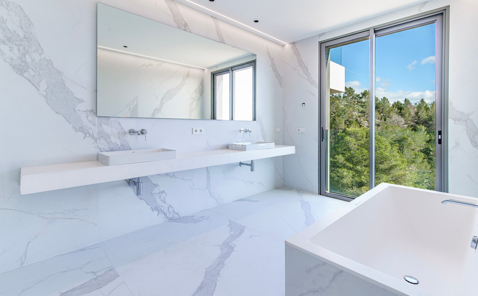 Baño principal luminoso con paredes y suelos de mármol blanco, lavabo doble, bañera y vistas al bosque.