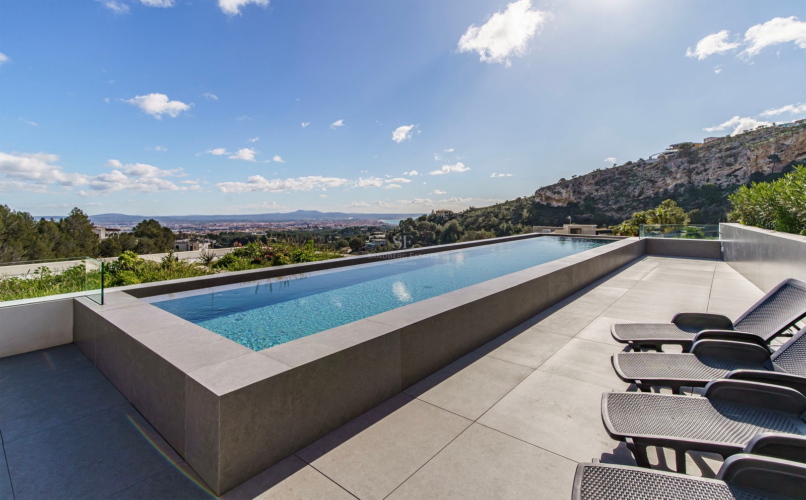 Piscina infinita en una terraza de piedra con tumbonas y vistas despejadas a la ciudad y la montaña.