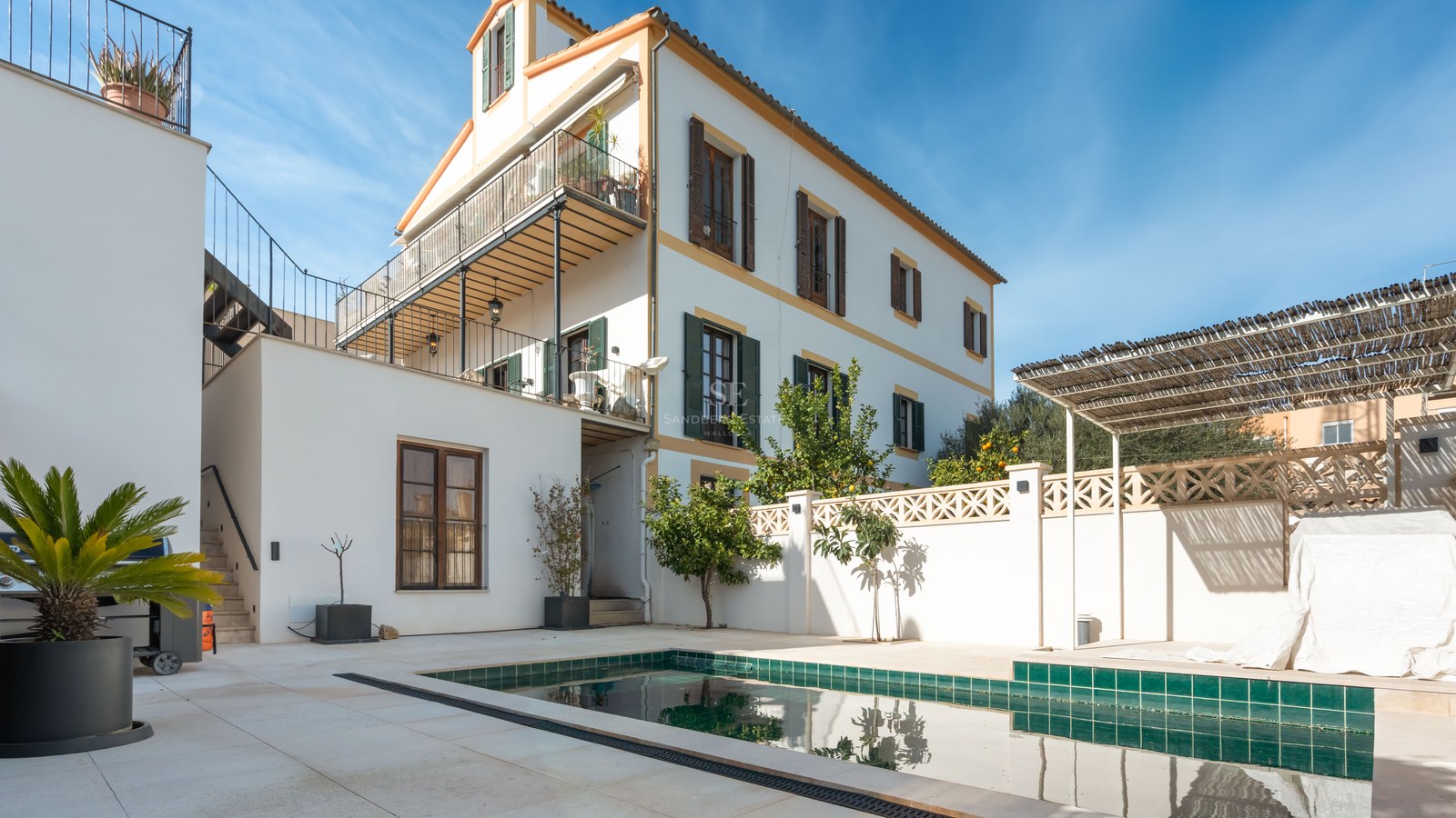 Grönkaklad pool på en vit stenterrass bredvid en traditionell villa med fönsterluckor i trä.