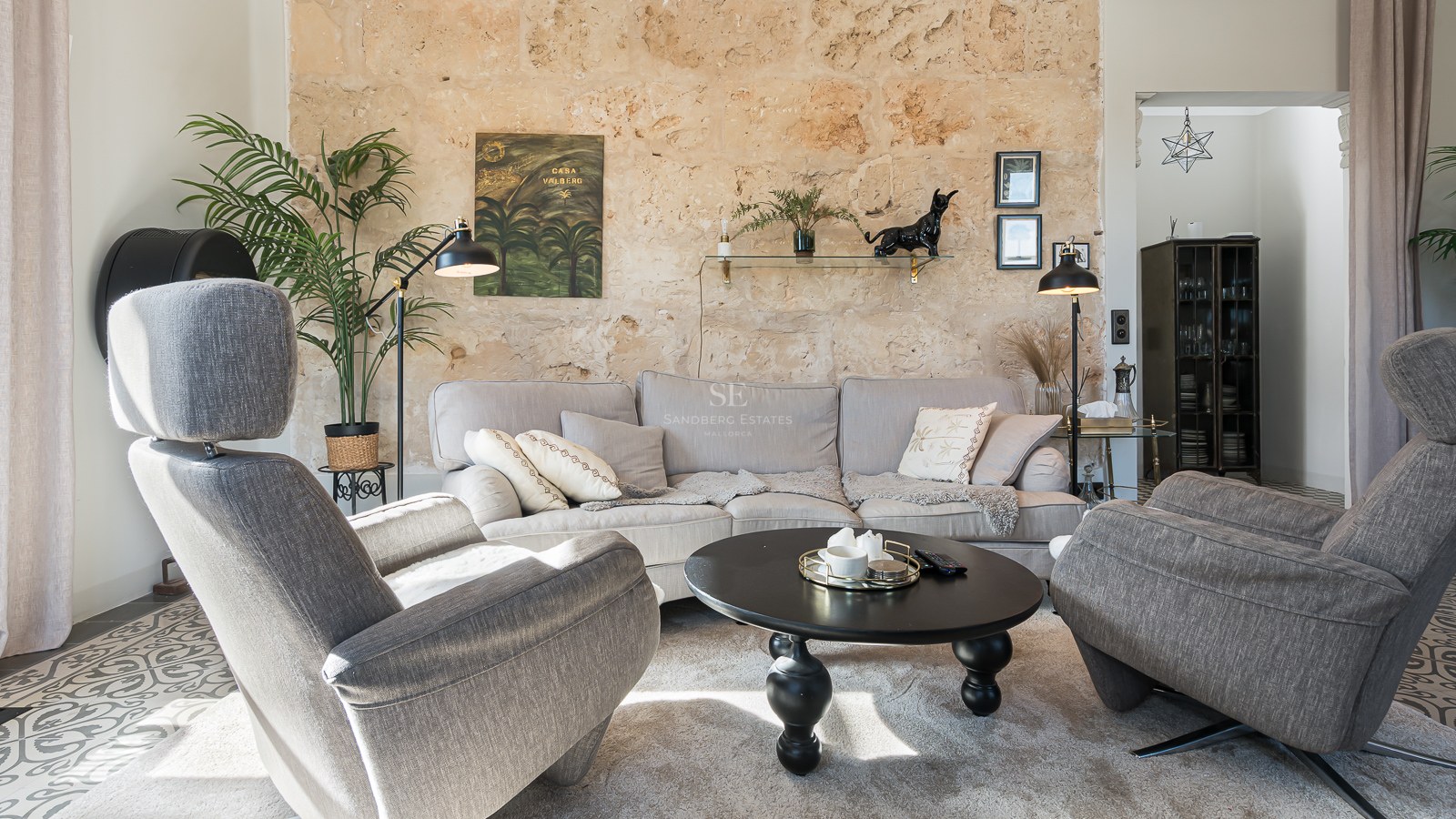 Modernt vardagsrum med grå fåtöljer, beige soffa, svart soffbord och en stor rustik stenvägg.