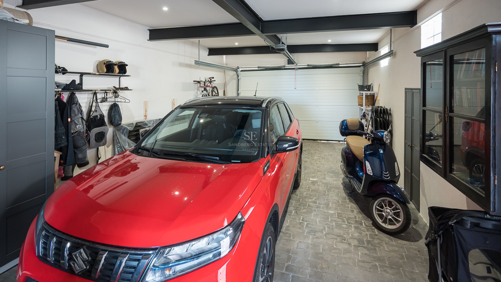 Ljust garage med röd bil, blå skoter, småstensgolv och svarta stålbalkar i taket.