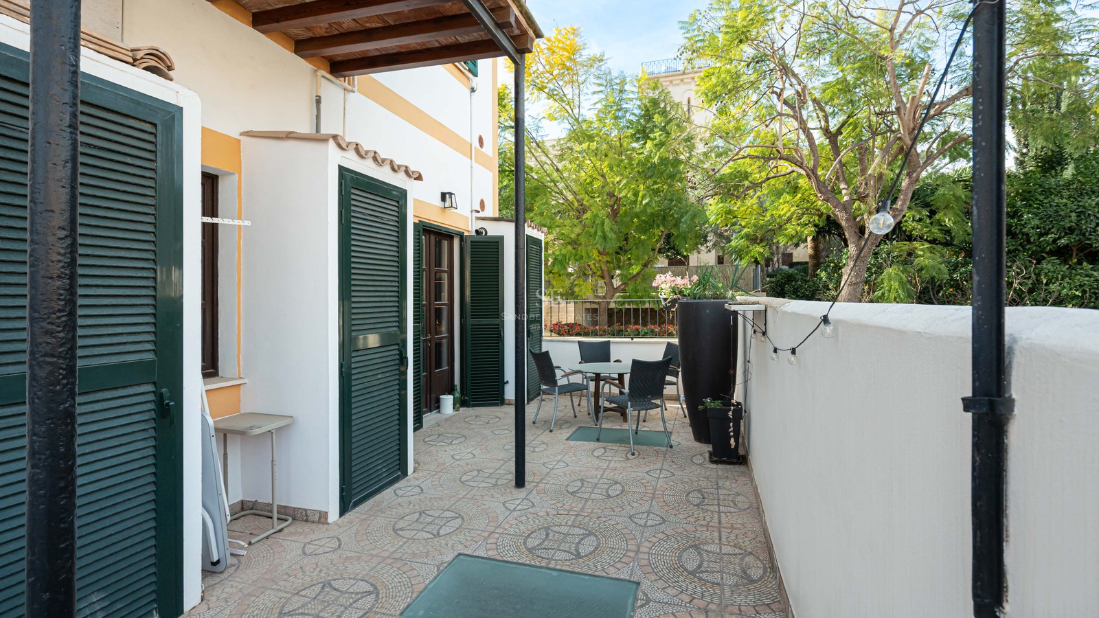 Utomhusterrass med bord, stolar, mönstrat klinkergolv och gröna fönsterluckor under en pergola.