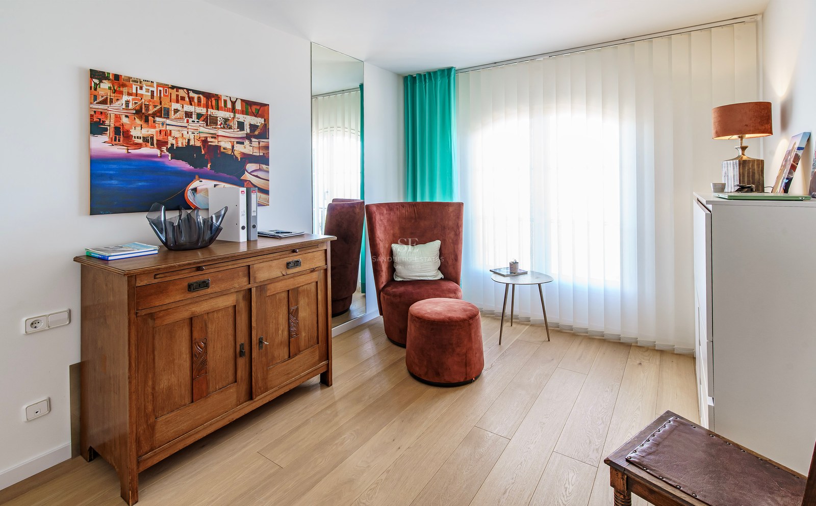 Cozy seating area with a terracotta velvet chair, wooden sideboard, and coastal artwork on light wood flooring.