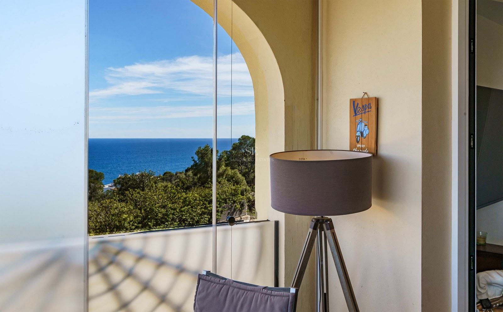 A covered terrace with frameless glass curtains looking out over pine trees to the blue Mediterranean sea.