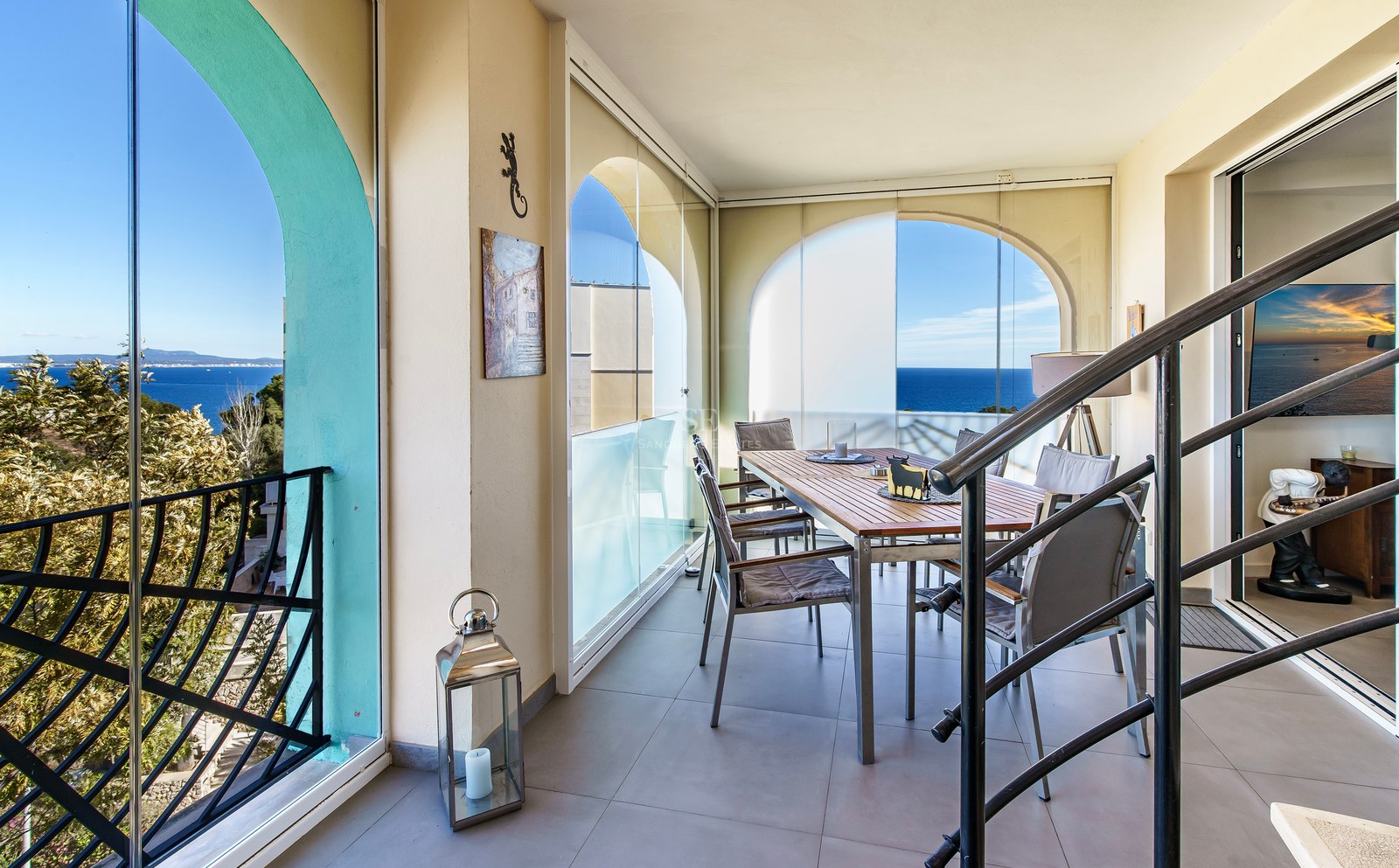 Covered dining terrace with wooden table, glass curtains, and views of the blue Mediterranean sea through arched openings.