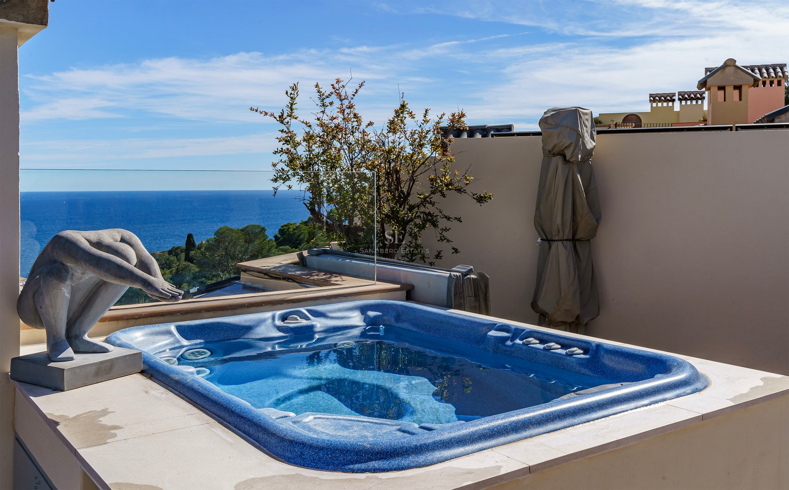 Blue built-in jacuzzi on a stone terrace with glass railings overlooking the Mediterranean Sea and a stone sculpture.