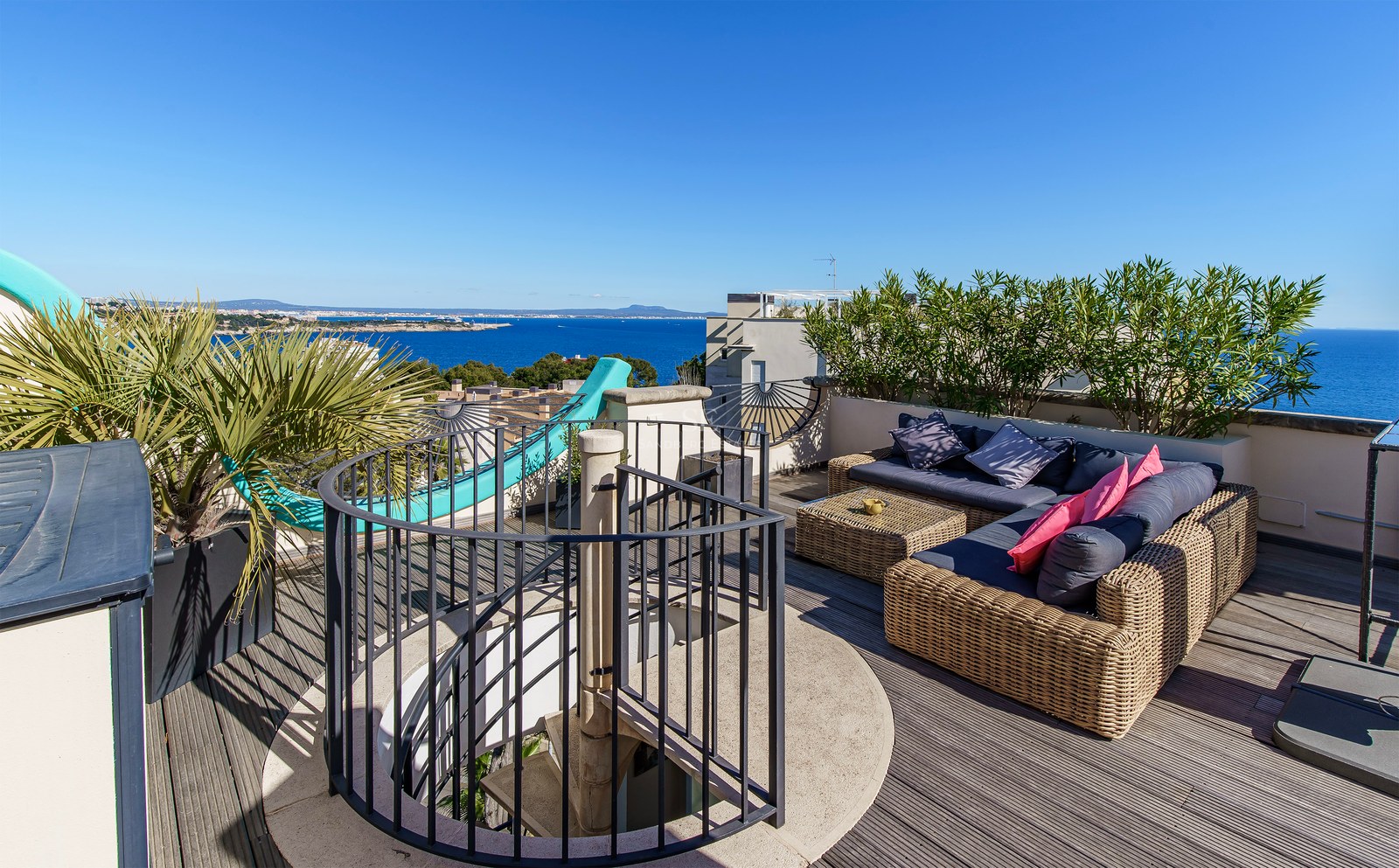 A sun-drenched wooden rooftop terrace with wicker sofas and a spiral staircase, overlooking the Mediterranean Sea.