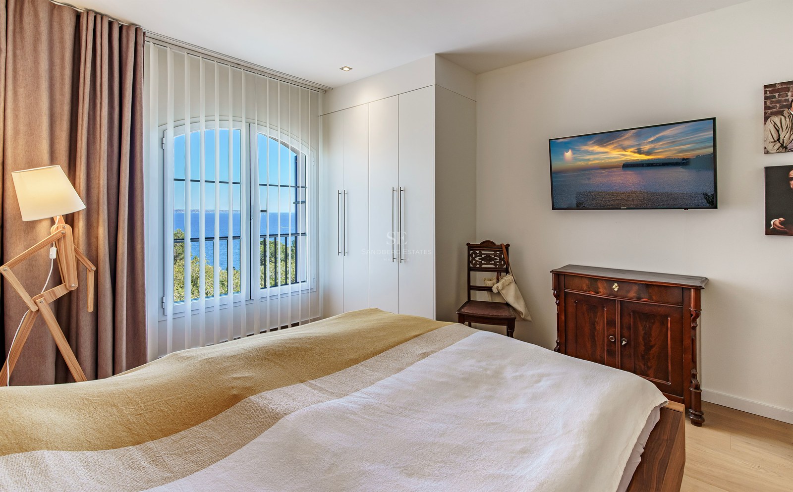 Bright bedroom with an arched window framing a Mediterranean sea view, featuring white wardrobes and wooden flooring.